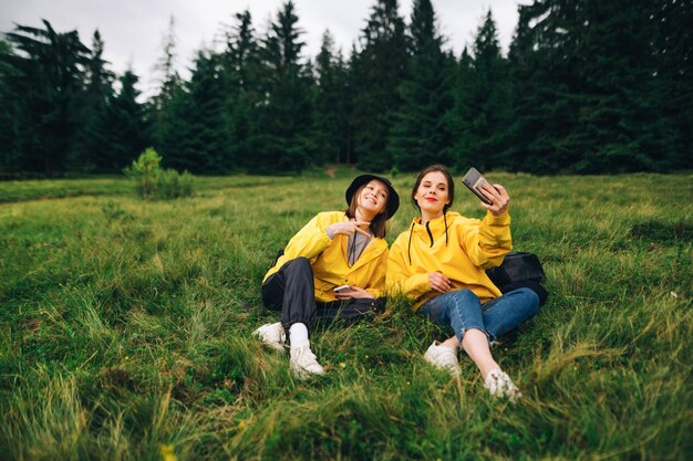 Dos turistas alegres están sentadas en un prado de montaña en una caminata y tomando selfies en una cámara de teléfono inteligente Mujeres atractivas con impermeables amarillos toman selfies descansando en la hierba en las montañas