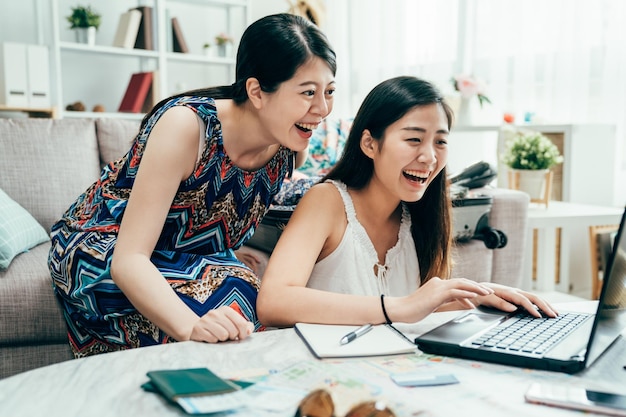 Dos turistas sorprendidas que encuentran ofertas en línea de viajes de vacaciones de verano en el sitio web usando una computadora portátil en el apartamento por la tarde. chicas jóvenes riendo alegre planificación ruta turística en el extranjero