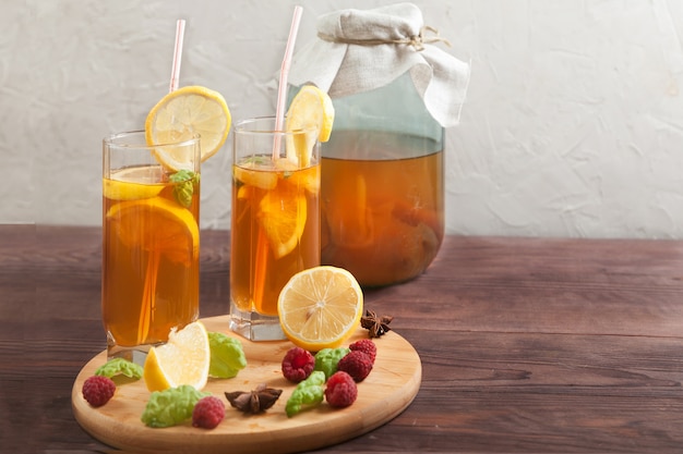 Dos vasos y banco con kombucha y rodajas de limón y frambuesas sobre una mesa de madera.