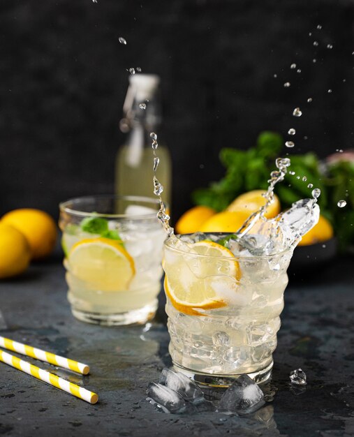Dos vasos de bebida de limonada con limón fresco y salpicaduras Cócteles refrescantes con limón, menta y hielo sobre fondo oscuro Concepto de bebidas frías de verano
