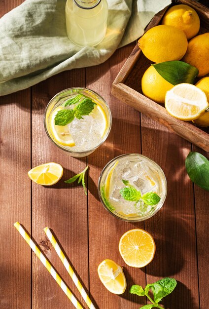 Dos vasos de bebida de limonada con limones frescos Cócteles refrescantes con menta de limón y hielo sobre fondo de madera oscura con sombras matutinas Concepto de bebidas frías de verano Vista superior