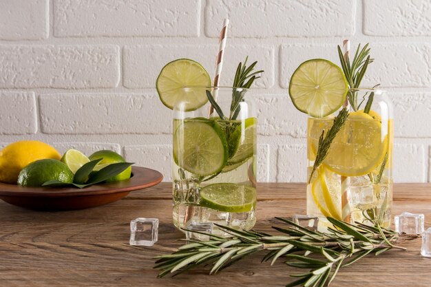 Dos vasos de bebidas refrescantes de verano y rodajas de limón y lima una ramita de llovizna y cubitos de hielo sobre una mesa de madera contra una pared de ladrillo blanco