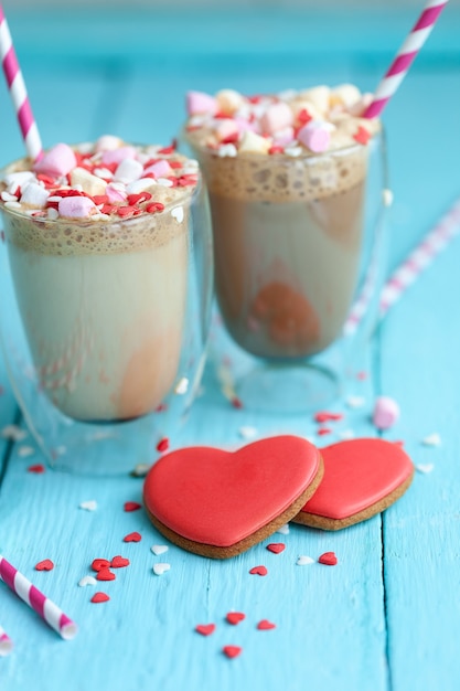 Dos vasos de café caliente o capuchino con malvaviscos y galletas en forma de corazón sobre un fondo de madera azul claro. Día de San Valentín de vacaciones.