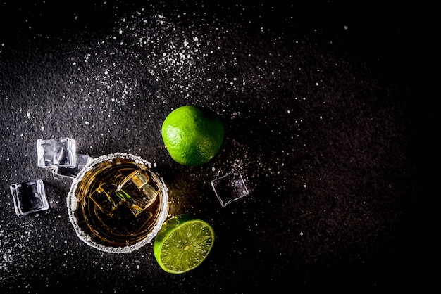 Dos vasos de chupito de tequila en la mesa oscura, con cubitos de hielo, sal y limas