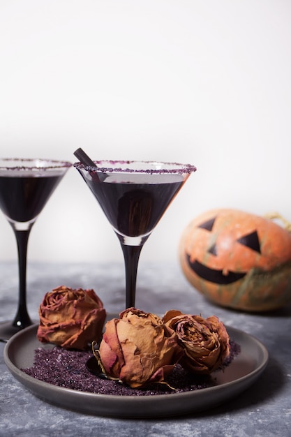 Dos vasos con cóctel negro, rosas secas, Jack-o'-lantern para la fiesta de Halloween en el fondo oscuro