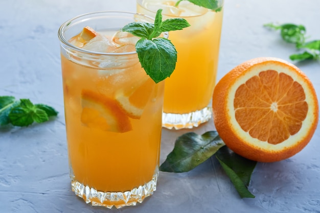 Dos vasos de jugo de naranja natural con rodajas de naranja, hielo, hojas de menta sobre una superficie gris