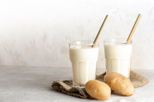 Dos vasos de leche de patata vegana con pajitas de bambú y tubérculos de patata sobre fondo neutro