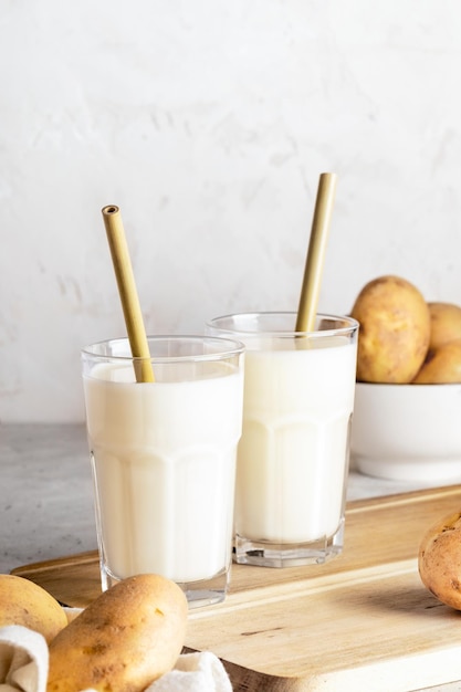 Dos vasos de leche de patata vegana y tubérculos de patata en tablero de cocina de madera sobre fondo blanco.