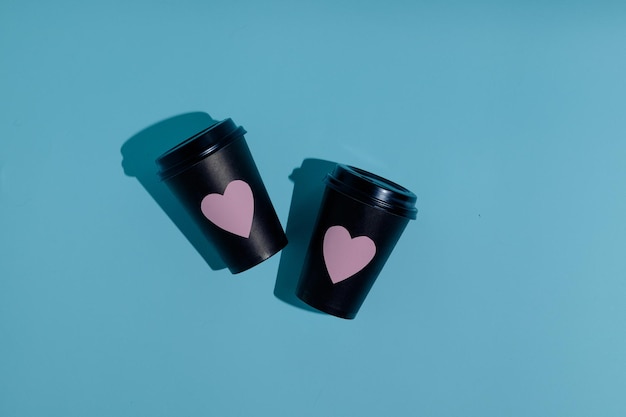 Dos vasos de papel negro para bebidas calientes con corazones rosas sobre fondo azul vacío vista superior plana