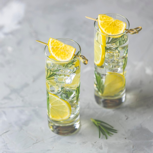 Dos vasos de refrescante bebida de lima limón con cubitos de hielo en copas de vidrio sobre un fondo gris claro Cóctel de refresco de limón fresco de verano con enfoque selectivo de romero