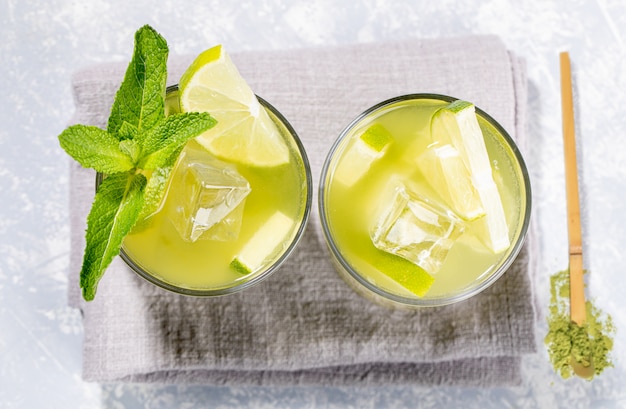 Dos vasos de té verde matcha helado con limón, hielo, menta y una cuchara de madera de polvo en gris. Vista superior.
