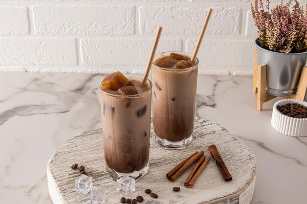 Dos vasos de vidrio con café con leche frío o con una bebida de chocolate y cubitos de hielo canela en una pizarra blanca de madera y una mesa de mármol