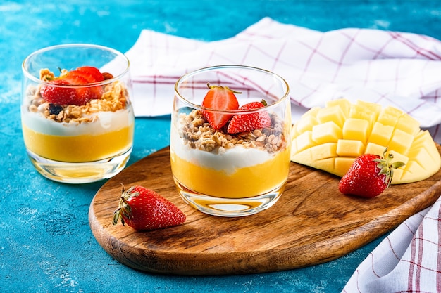 Dos vasos de vidrio con postre de frutas de mango y fresa con muesli y crema o ricotta sobre tabla de madera