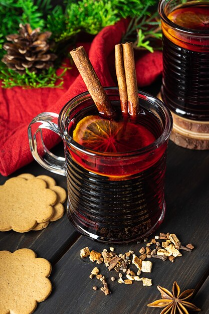 Dos vasos de vino tinto caliente caliente con canela y limón. Especias, galletas de jengibre, rama de abeto y cono. Fondo de madera oscura. Ambiente cálido navideño.