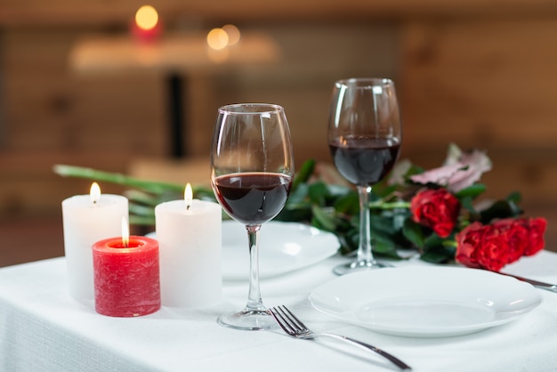 Dos vasos con vino tinto, velas encendidas y un ramo de rosas en un primer plano de mesa servido en el interior.