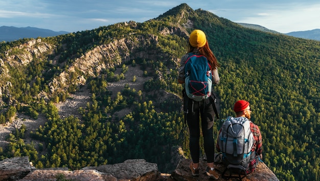 Dos viajeros en el fondo de las montañas, panorama. Viajeros al atardecer en las montañas. Turistas con mochilas al borde del acantilado. Una pareja admira la puesta de sol desde un acantilado. Copia espacio