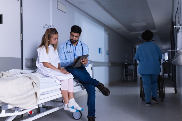 Foto doutor diversificado a falar com uma rapariga a usar um tablet na cama no corredor do hospital