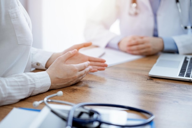 Doutor e paciente sentados e discutindo algo em uma mesa de madeira enquanto usam um computador portátil. O foco é nas mãos do paciente. Conceito de medicina.