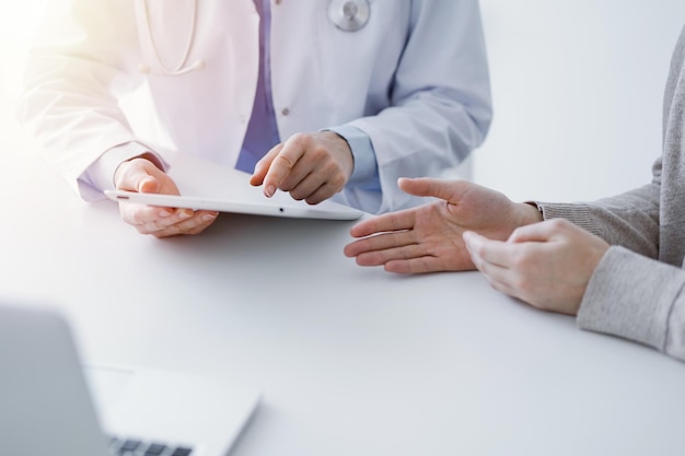 Doutor e paciente sentados na mesa na clínica. O foco é nas mãos da médica usando computador tablet, em close-up. Conceito de medicina e cuidados de saúde