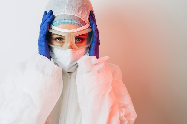 Doutor em uniforme de proteção, óculos, luvas, sobre um fundo azul na luz de neon. retrato de close-up de um médico em néon vermelho. homem cansado está lutando contra um coronavírus. COVID 19