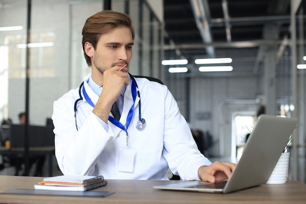 Doutor em uniforme médico branco com estatoscópio, lendo o arquivo pessoal do paciente no tablet e grava dados no laptop.