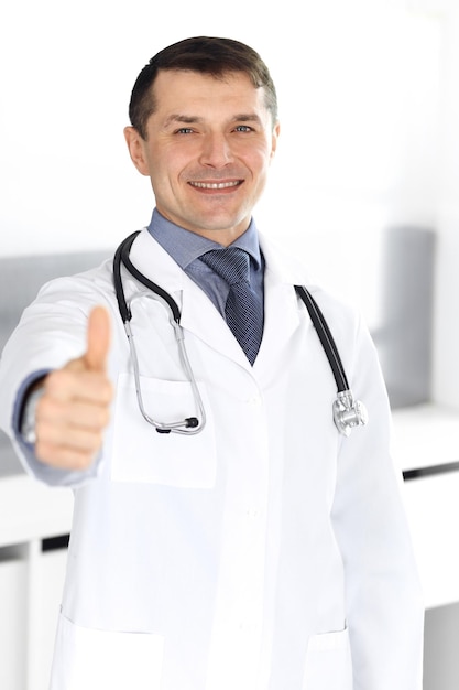 Doutor homem alegre sorrindo para a câmera, polegares para cima. serviço médico perfeito na clínica. futuro feliz em medicina e saúde.