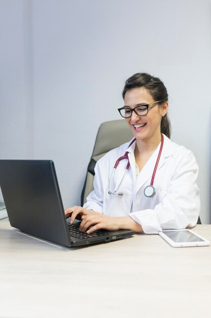 Foto doutor sorridente usando laptop enquanto está sentado na mesa na clínica