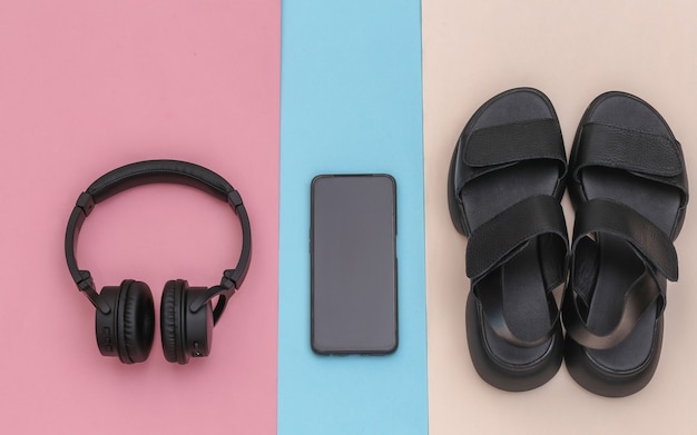 Drahtlose Stereokopfhörer und Smartphone, Sandalen auf rosa blauem Pastellhintergrund. Ansicht von oben