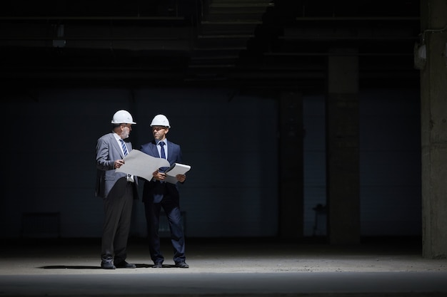 Dramático retrato de cuerpo entero de dos empresarios maduros vistiendo cascos y sosteniendo planes mientras está de pie en la oscuridad en el sitio de construcción iluminado por una iluminación intensa,