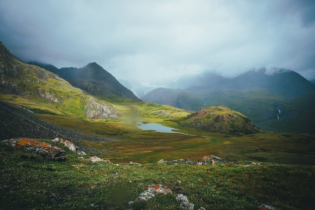 Dramatische Alpenlandschaft mit wunderschönem Bergsee im grünen Tal zwischen großen Felsen und hohen Bergen bei bedecktem Wetter. Tolle Aussicht auf See und Gletscher in niedrigen Wolken. Stimmungsvolle Landschaft.