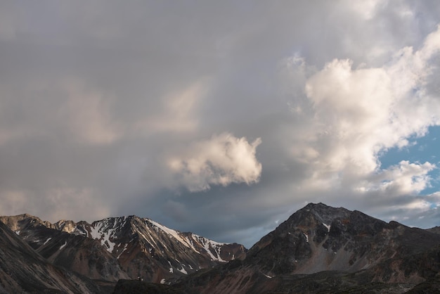 Dramatische Berglandschaft mit hoher Bergkette mit scharfem Felsgipfel unter Wolken in Sonnenuntergangsfarbe am düsteren Himmel. Dunkle atmosphärische Landschaft mit großen Bergen mit Schnee bei wechselhaftem Wetter