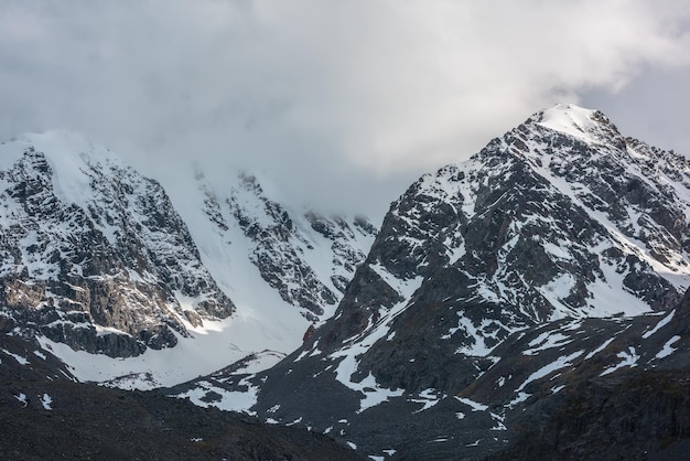 Dramatische Landschaft mit sonnenbeschienener, schneebedeckter Bergkette bei tief bewölktem Himmel. Goldener Glanz auf weißem Schnee und schwarzen Felsen in tief hängenden Wolken. Fantastischer Blick auf schneebedeckte Berge mit goldenem Sonnenlicht bei wechselhaftem Wetter