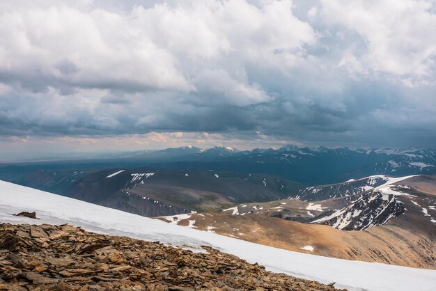 Dramatischer Alpenblick vom Steinhügel mit Schnee bis zum schneebedeckten Berggipfel im Licht unter bewölktem Himmel. Stimmungsvolle Landschaft in großer Höhe mit Bewölkung. Hoher schneebedeckter Berggipfel im Sonnenlicht bei bewölktem Himmel