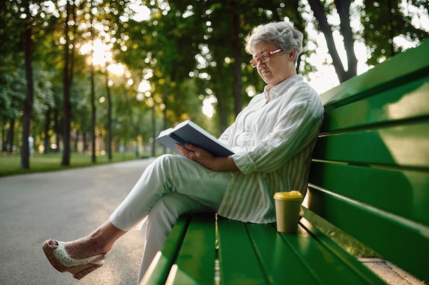 Dranny de óculos, lendo um livro no banco do parque de verão. Estilo de vida de pessoas idosas. Vovó bonita se divertindo ao ar livre, idosa na natureza