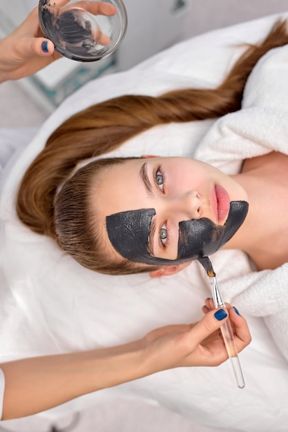 Draufsicht auf beschnittener professioneller Kosmetiker, der schwarze Maske auf frisches Gesicht einer schönen entspannten kaukasischen Frau für Carbon-Peeling anwendet. Hautpflege, Wohlbefinden, Spa, Beauty-Konzept. Platz kopieren