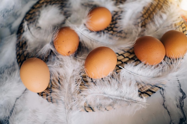 Draufsicht auf braune Hühnereier und flauschige Vogelfedern auf Korbständer auf Marmortisch. Rustikale Komposition mit österlicher Atmosphäre. Konzept von landwirtschaftlichen Produkten, gesundem Essen, Osterferien.