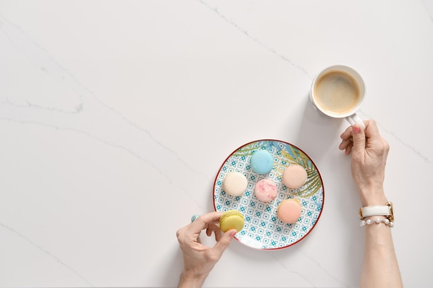 Draufsicht auf bunte Kuchenmakronen auf einem Teller und weibliche Hand mit einer Tasse Kaffee