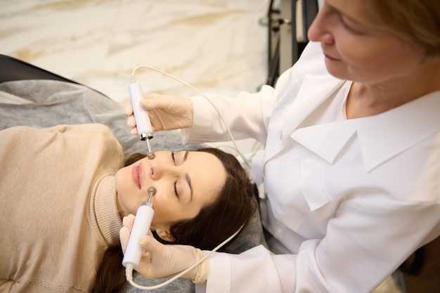 Draufsicht auf das weibliche Gesicht während der Mikrostromtherapie Schöne Europäerin in der Spa-Klinik, die eine anregende elektrische Gesichtsbehandlung von einem Schönheitstherapeuten erhält