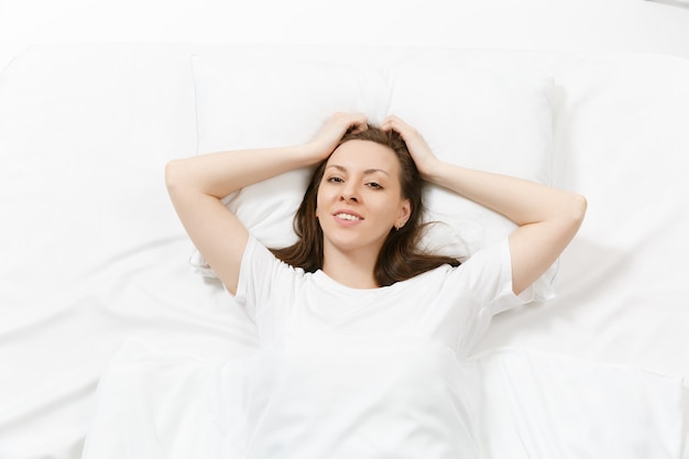 Draufsicht auf den Kopf einer glücklichen brünetten jungen Frau, die mit weißem Laken, Kissen, Decke im Bett liegt