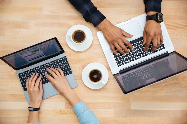 Draufsicht auf die Hände eines afrikanischen Mannes und einer kaukasischen Frau, die auf zwei Laptops tippen und Kaffee auf einem Holztisch trinken