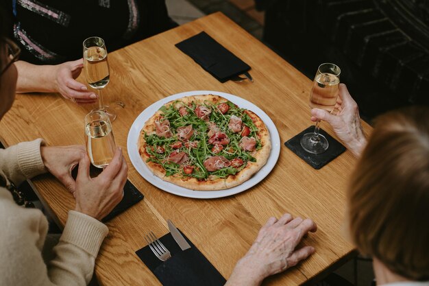 Draufsicht auf die Hände nicht erkennbarer älterer Menschen, möglicherweise Frauen, die Pizza essen und Wein oder Champagner in einem gemütlichen Café im Freien trinken, um ihr Treffen oder einen Urlaub zu feiern