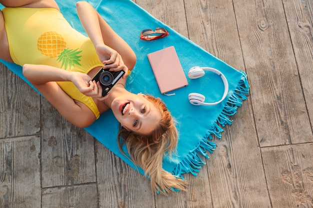 Draufsicht auf ein attraktives lächelndes junges blondes Mädchen mit Badeanzug, das sich auf einer Sommerdecke am Strand über Holzboden entspannt und Fotokamera hält