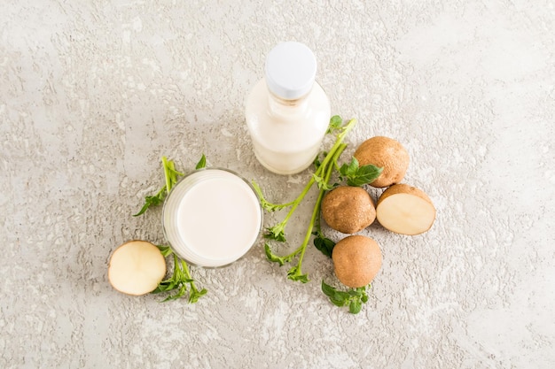 Draufsicht auf ein mit Kartoffelmilch gefülltes Glas eine Flasche Pflanzenmilch und Kartoffelknollen grauer Betonhintergrund