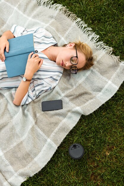 Draufsicht auf ein schönes junges blondes Mädchen, das sich auf einem Rasen im Park entspannt und ein Buch hält, während es mit dem Handy auf einer Decke liegt