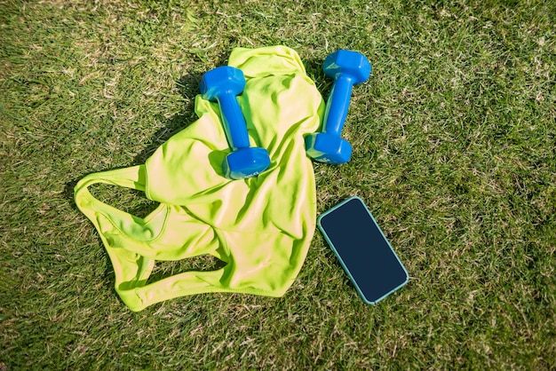 Draufsicht auf ein Sporttrikot mit Hanteln und einem Smartphone auf dem Rasen eines Fußballfeldes