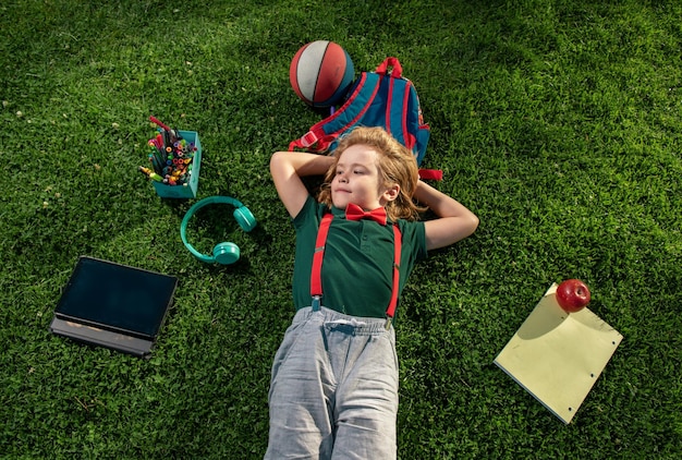 Draufsicht auf ein Vorschulschülerkind, das auf dem Gras liegt und im Freien einen Unterricht nimmt, ein glücklicher Junge, der sich entspannt