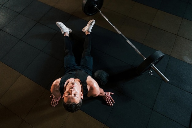 Draufsicht auf einen starken Mann in sportlicher Kleidung, der auf dem Boden sitzt und eine Pause im Fitnessstudio macht