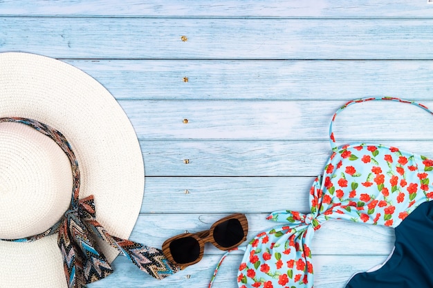 Draufsicht auf einen strohweißen Hut mit Brille und Badeanzug, auf blauem Holzhintergrund liegend. Sommerferienkonzept
