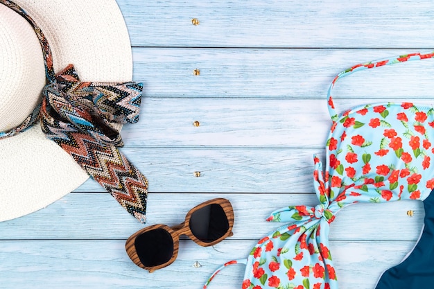 Draufsicht auf einen weißen Strohhut mit Brille und Badeanzug, auf blauem Holzhintergrund liegend. Sommerferienkonzept.