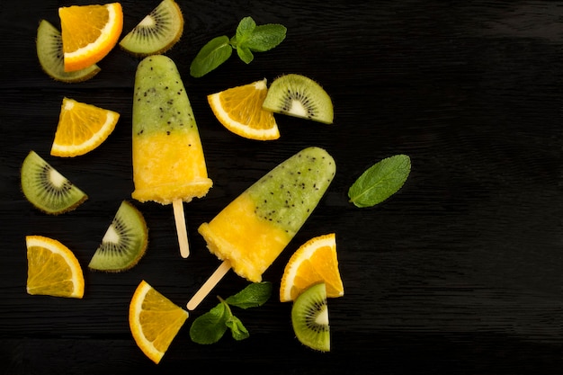 Draufsicht auf Eis am Stiel mit Kiwi und Orangenfrucht auf dem schwarzen Holztisch. Speicherplatz kopieren.